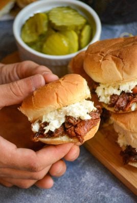 a hand holding up a coleslaw and pickle topped pulled pork slider