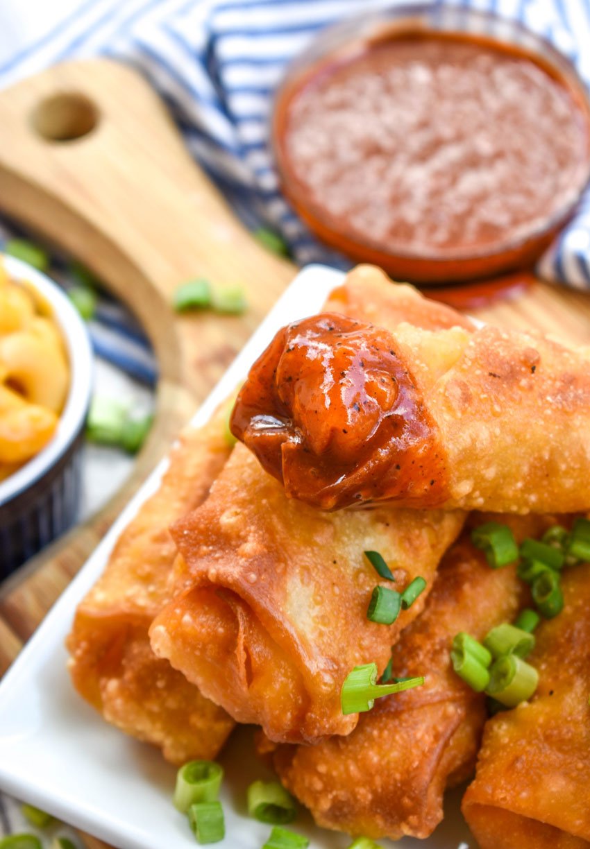 a mac and cheese egg roll dipped in barbecue sauce resting on a stack of egg roll son a white platter