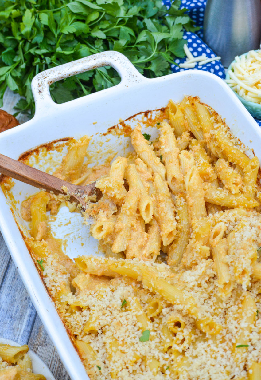 a wooden spoon digging into a white casserole dish filled with pumpkin macaroni and cheese with white cheddar