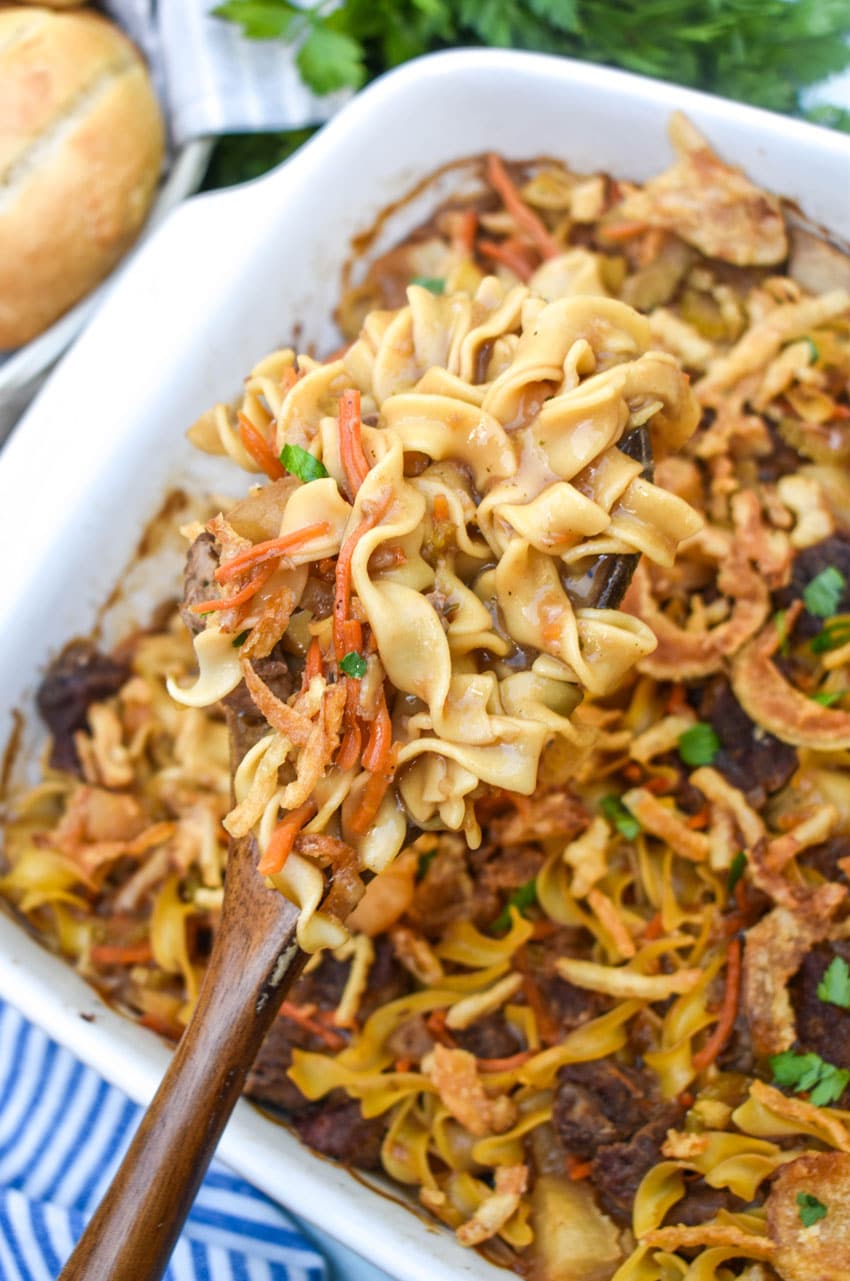 a wooden spoon scooping beef stew noodle casserole out of a large white baking dish