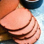 a chub of smoked bologna shown sliced on a wooden cutting board with barbecue sauce & mustard in the background