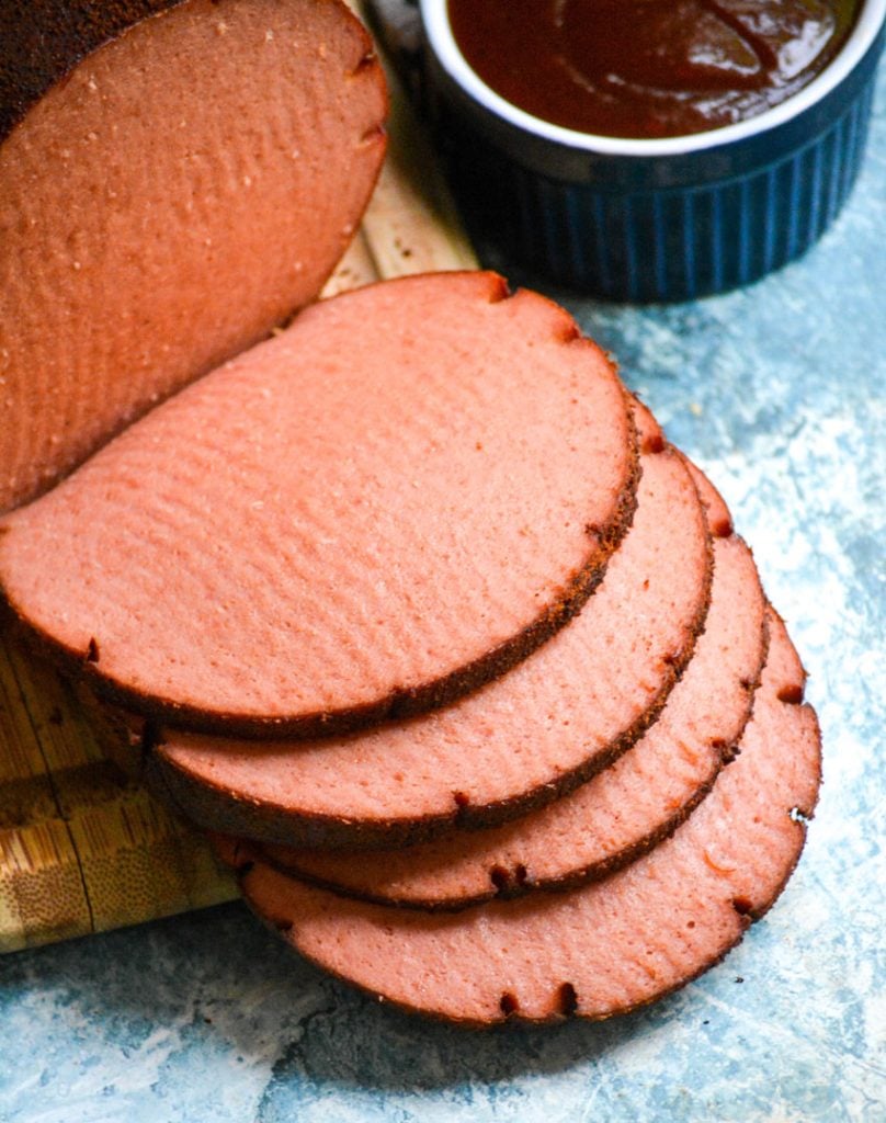a chub of smoked bologna shown sliced on a wooden cutting board with barbecue sauce & mustard in the background