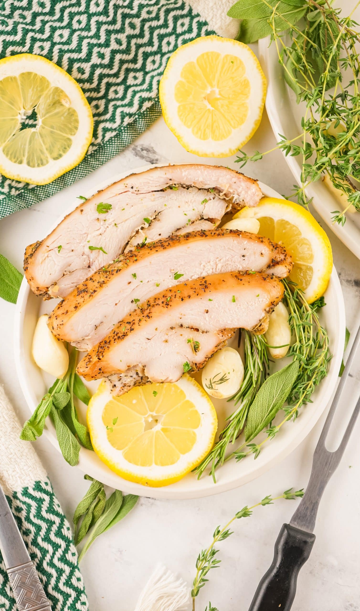 slices of a smoked turkey breast on a bed of fresh herbs and sliced lemons on a white platter