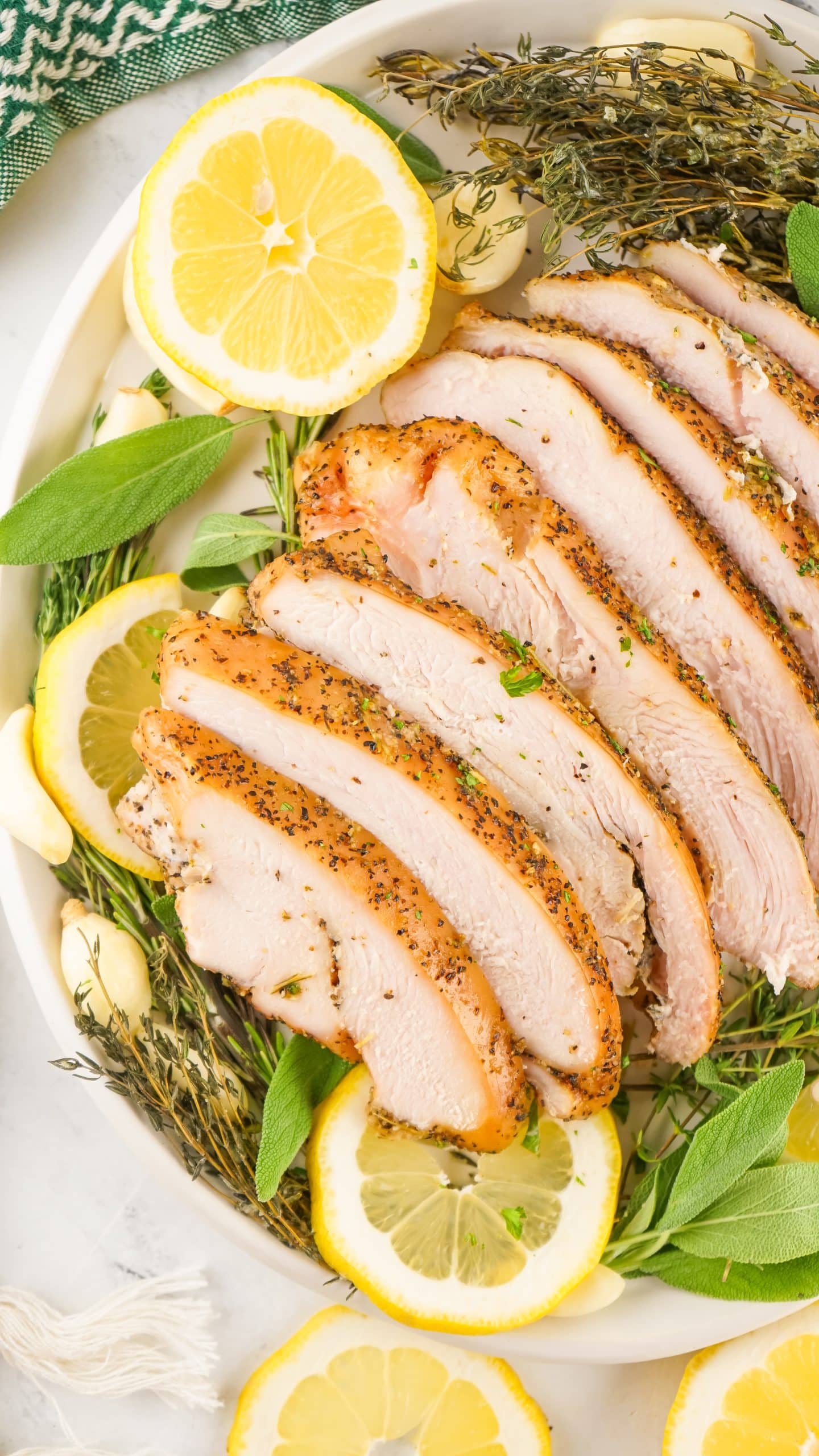 slices of a smoked turkey breast on a bed of fresh herbs and sliced lemons on a white platter